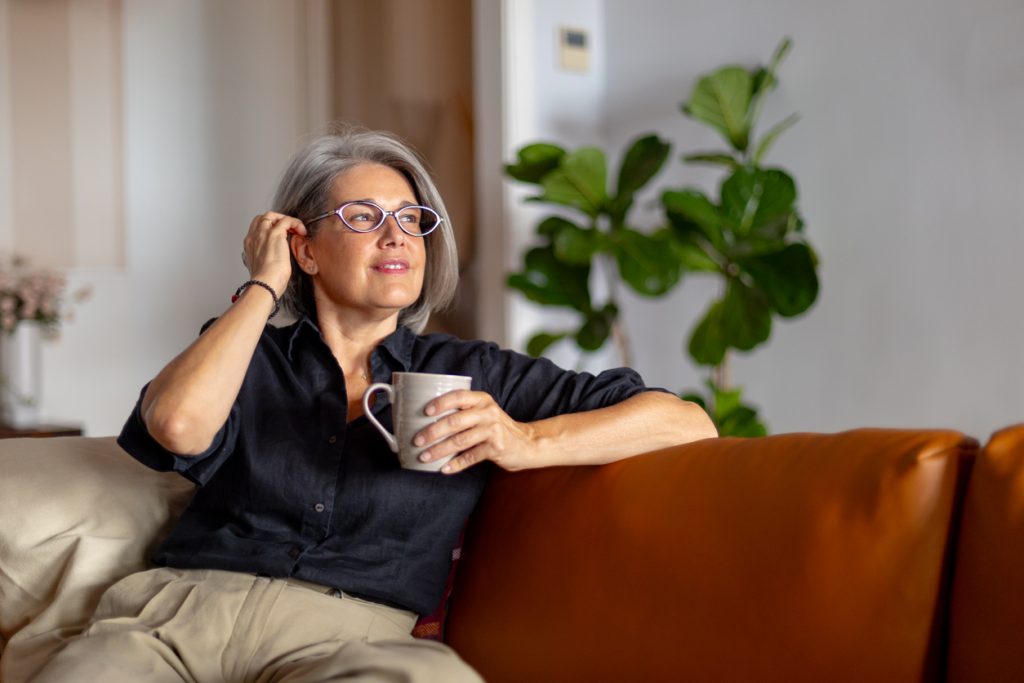 Uma mulher de cabelos grisalhos e óculos, sentada pensativamente em um sofá de couro marrom. Ela segura uma caneca cinza e coloca a mão no cabelo, refletindo sobre as mudanças internas da menopausa.