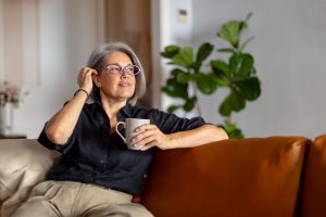 Uma mulher de cabelos grisalhos e óculos, sentada pensativamente em um sofá de couro marrom. Ela segura uma caneca cinza e coloca a mão no cabelo, refletindo sobre as mudanças internas da menopausa.
