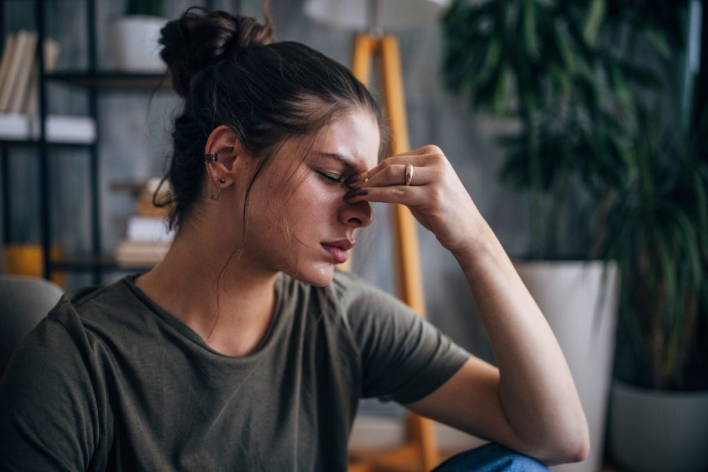 Uma mulher jovem sentada em um ambiente interno, com os olhos fechados e apertando a ponte do nariz com a mão, demonstrando uma fisionomia de tensão, cansaço ou dor de cabeça. A imagem ilustra como o estresse físico e mental é um dos fatores que podem causar glicose alta.