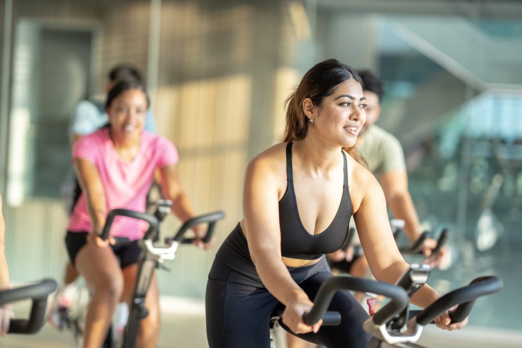 Fotografia de uma mulher jovem sorridente, vestindo top preto de ginástica, pedalando ativamente em uma bicicleta ergométrica durante uma aula em ambiente claro. Ao fundo, outras pessoas desfocadas também praticam o exercício. A cena representa a prática de atividades físicas essenciais para vencer o sedentarismo e potencializar o emagrecimento.