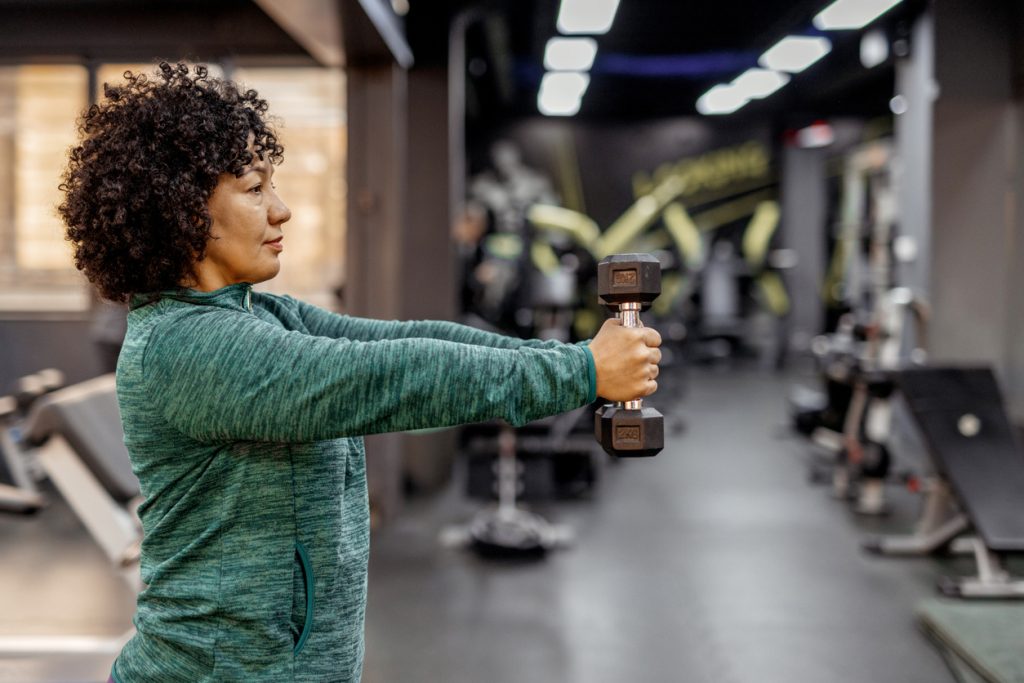 Mulher adulta com cabelos cacheados segurando um halter à frente do corpo em uma academia. A imagem ilustra o artigo principal que explica por que a musculação emagrece e é aliada na perda de peso.