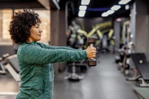 Mulher adulta com cabelos cacheados segurando um halter à frente do corpo em uma academia. A imagem ilustra o artigo principal que explica por que a musculação emagrece e é aliada na perda de peso.