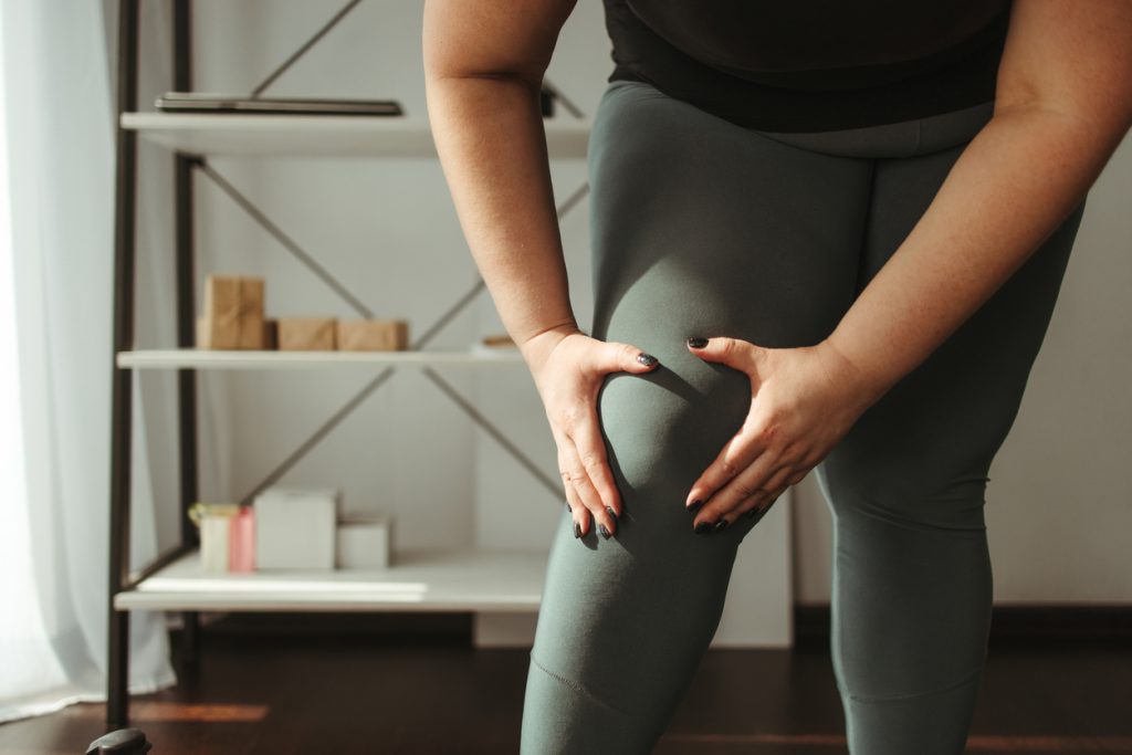Mulher com sobrepeso em pé, vestindo roupas confortáveis verdes, segurando e massageando o joelho direito com as duas mãos, demonstrando desconforto e dor nas articulações, um sintoma comum da osteoartrite.