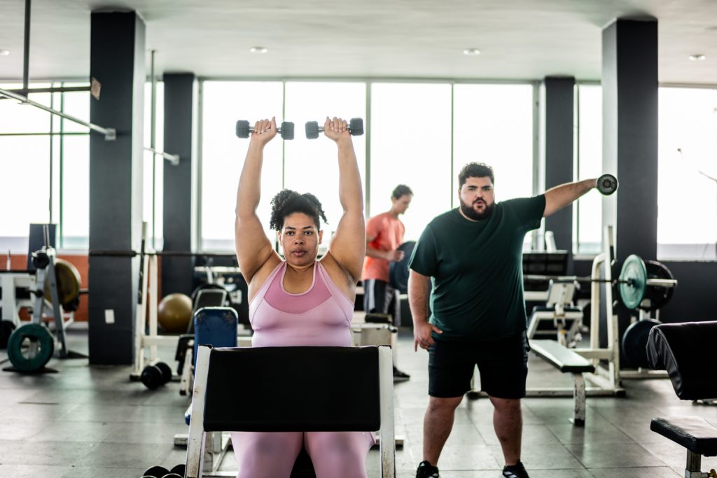 Mulher sentada em um banco de academia levantando dois halteres acima da cabeça, com um homem treinando ao fundo. A prática de exercícios de força comprova que a musculação emagrece e transforma a composição corporal de forma saudável.