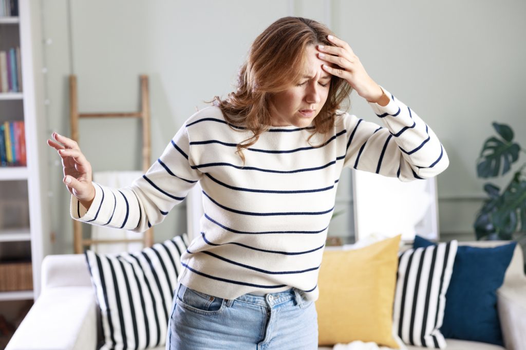 Mulher em pé na sala de casa, vestindo blusa listrada, com uma das mãos na testa e a outra estendida para o lado, expressando dor, confusão e desequilíbrio, ilustrando como identificar os primeiros sintomas de AVC.