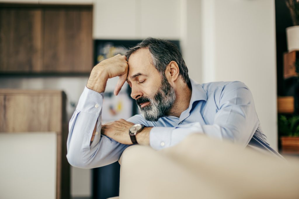 Homem de meia-idade vestindo camisa social azul, sentado no sofá com os olhos fechados e a mão apoiada na testa. Ele aparenta exaustão física e mental, ilustrando os sintomas e sinais de alerta da baixa testosterona após os 40 anos.