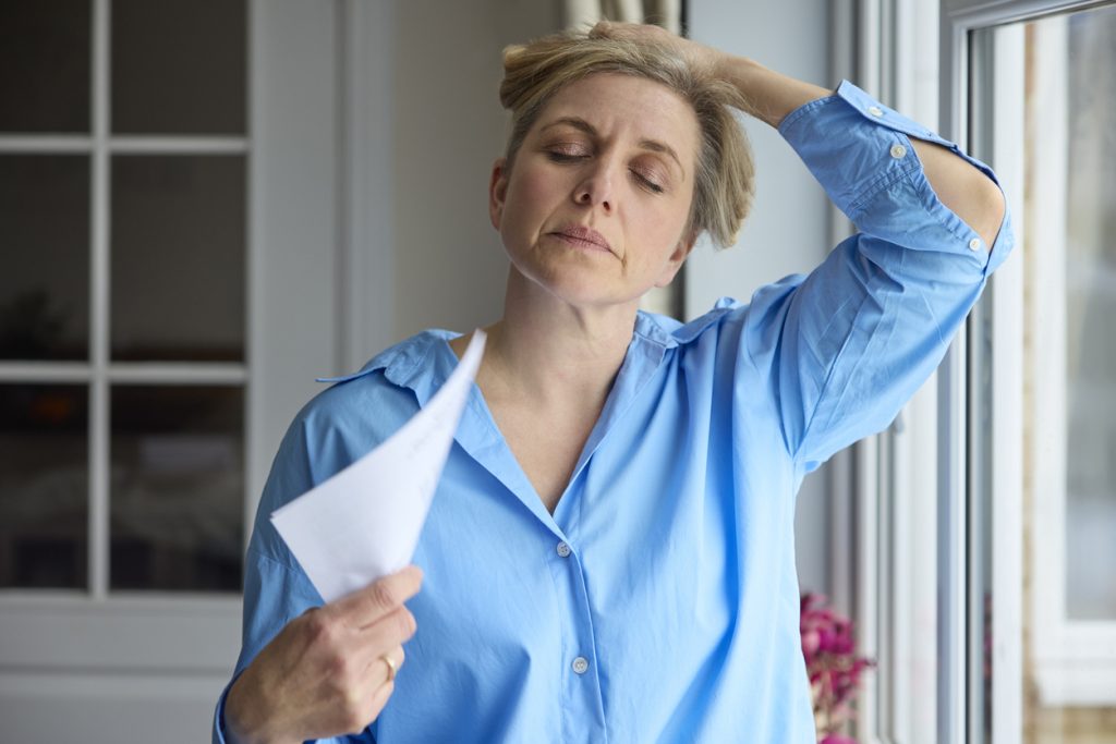 Uma mulher madura com expressão de desconforto por calor e cansaço, com a mão na cabeça e segurando papéis, em frente a uma janela. Esta imagem ilustra os sintomas difíceis que podem surgir na menopausa.