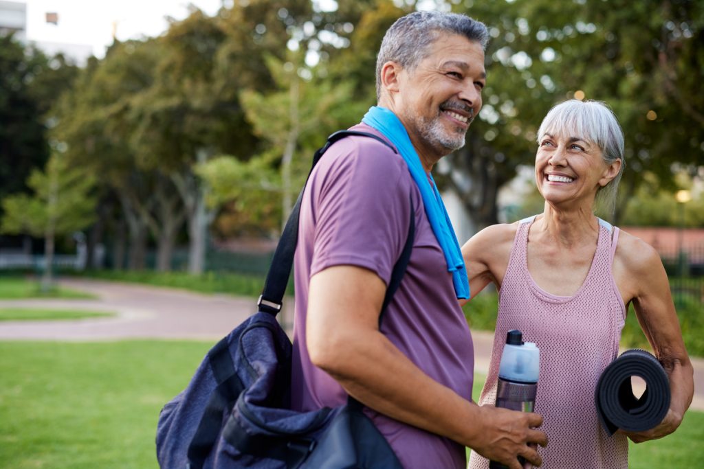 Casal mais velho sorrindo um para o outro em um ambiente ao ar livre. O homem carrega uma bolsa de academia no ombro e a mulher segura um tapete de yoga e uma garrafa de água, representando o companheirismo e a adoção de hábitos saudáveis na vida do casal durante a andropausa.