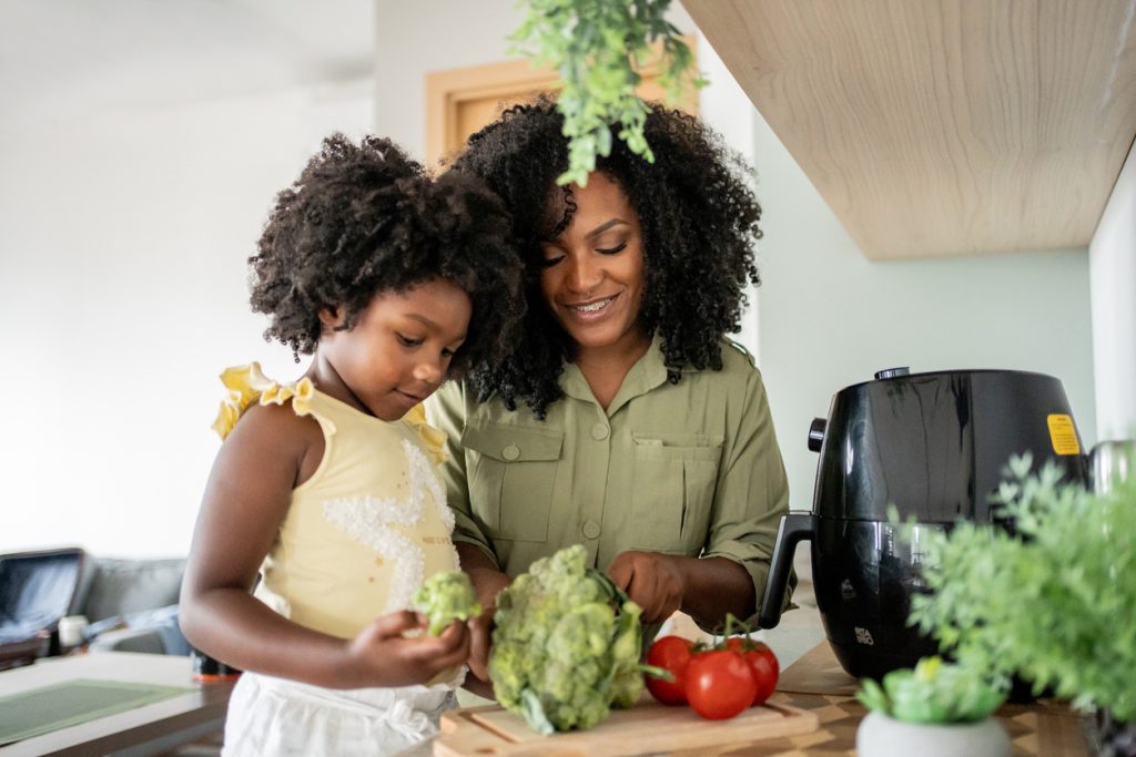 Mãe e filha negra sorrindo enquanto preparam vegetais frescos, como brócolis e tomates, na bancada da cozinha.