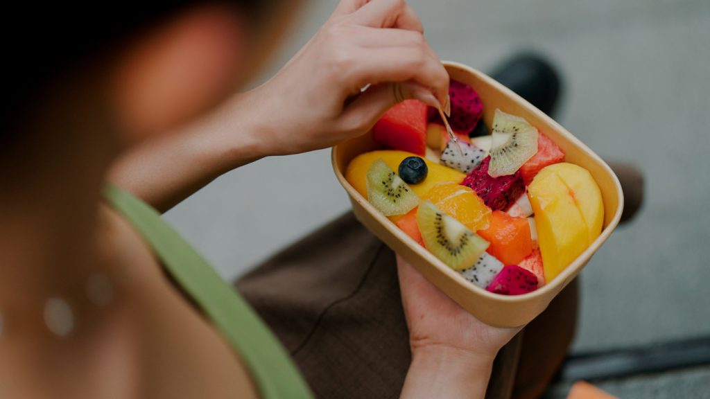 Pessoa segurando uma tigela de papelão cheia de frutas frescas picadas, como melancia, kiwi, manga e pitaya, ilustrando boas escolhas alimentares ricas em fibras para ajudar na constipação.