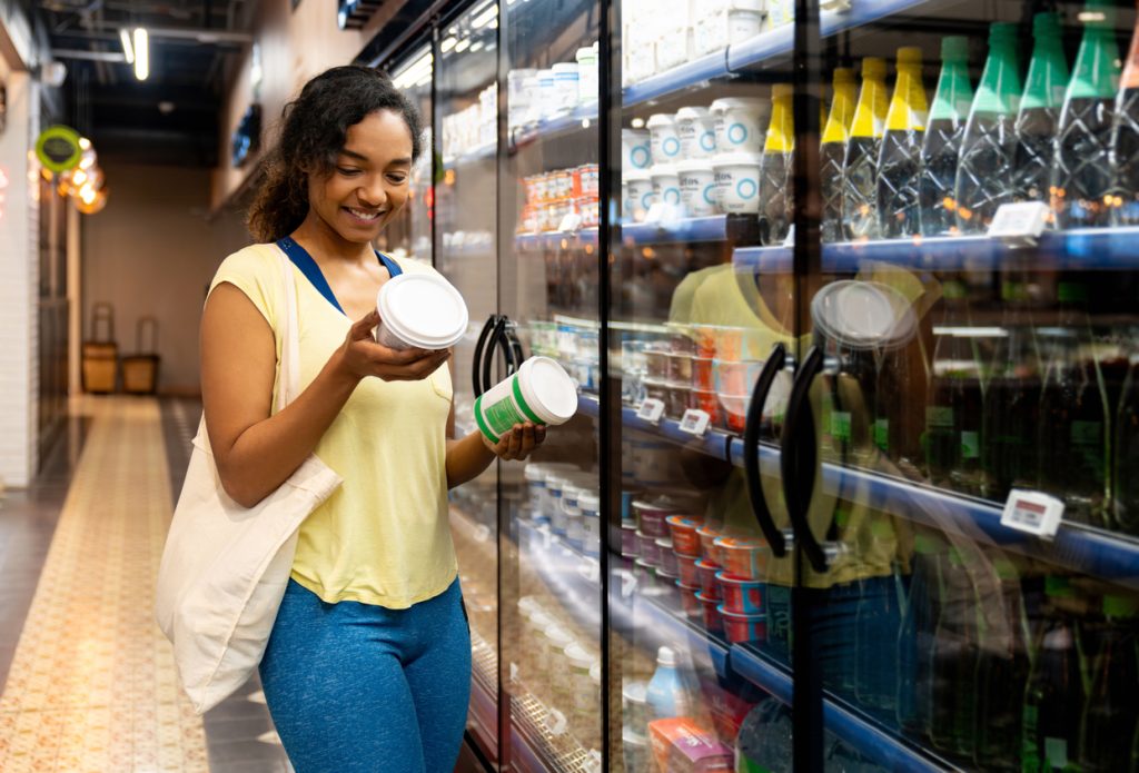 Mulher sorridente no corredor do supermercado segurando e lendo o rótulo de um produto refrigerado, refletindo o novo comportamento do consumidor na busca por bater metas de proteína.