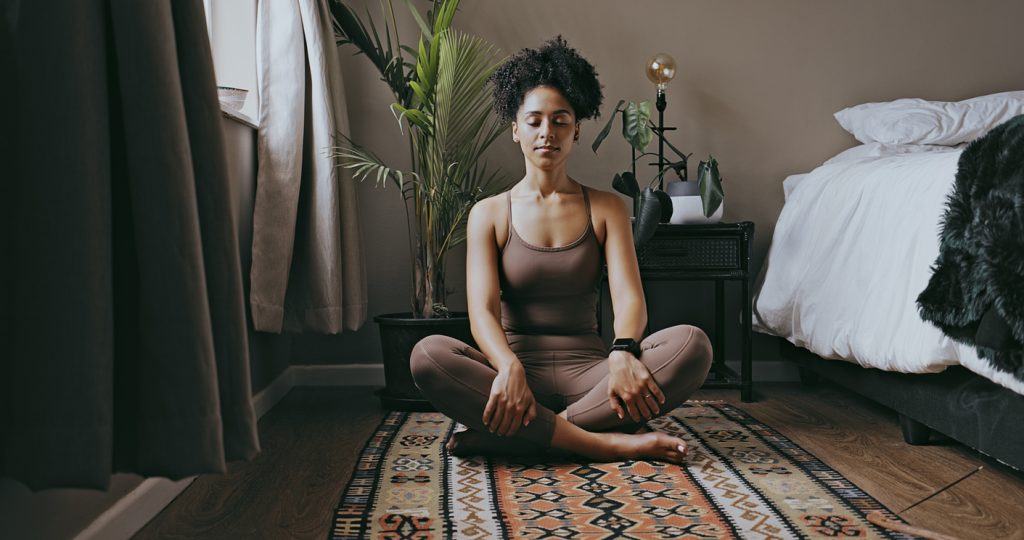 Mulher com roupas confortáveis sentada de pernas cruzadas em um tapete no quarto, meditando com os olhos fechados e expressão serena para relaxar a mente e combater a insônia. Ao fundo, há uma cama e uma planta.