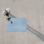 Vista aérea de uma mulher olhando para o próprio reflexo em um espelho retangular deitado no chão de concreto. O espelho reflete o céu azul em vez de um ângulo direto, ilustrando o conceito de distorção de imagem, onde a percepção da mente não corresponde à realidade física.