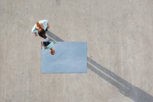 Vista aérea de uma mulher olhando para o próprio reflexo em um espelho retangular deitado no chão de concreto. O espelho reflete o céu azul em vez de um ângulo direto, ilustrando o conceito de distorção de imagem, onde a percepção da mente não corresponde à realidade física.