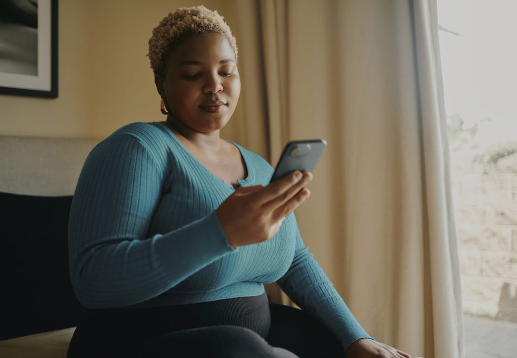 Mulher sentada em um ambiente interno, olhando com uma expressão séria e reflexiva para a tela do celular. O uso da internet e as comparações online são causas frequentes que despertam ou agravam a distorção de imagem.
