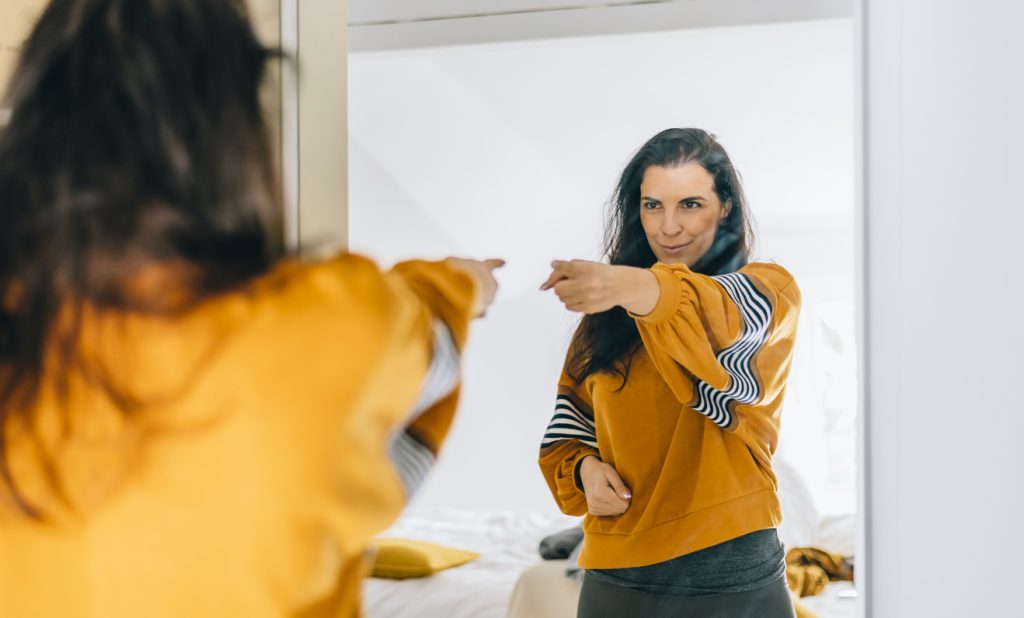 Mulher de blusa amarela sorrindo de forma confiante enquanto aponta de forma positiva para o seu próprio reflexo no espelho. A imagem transmite autoconfiança e ilustra o processo de cuidar da mente para superar a distorção de imagem e criar uma relação saudável com o corpo.