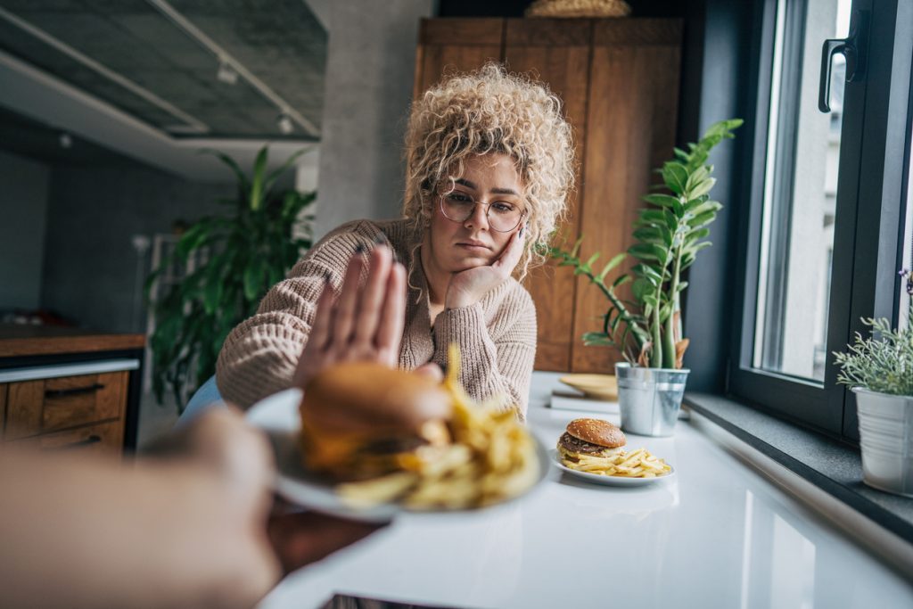 Mulher com óculos e cabelos cacheados loiros com expressão de aversão e desinteresse. Ela faz um gesto de "pare" com a mão, rejeitando um prato com hambúrguer e batatas fritas que está sendo oferecido a ela, representando os prejuízos à saúde causados pela falta de fome extrema.
