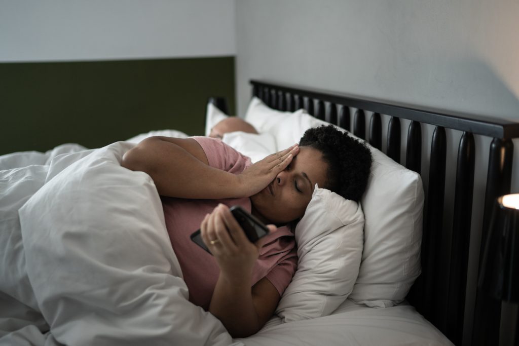 Mulher deitada na cama durante a noite com expressão de cansaço, esfregando o rosto enquanto segura um smartphone, ilustrando os efeitos da insônia. Ao fundo, um homem dorme.