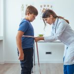 Menino em pé no consultório sendo medido na cintura com uma fita métrica por uma médica vestindo jaleco branco.