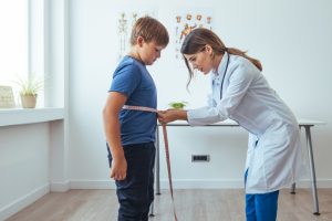 Menino em pé no consultório sendo medido na cintura com uma fita métrica por uma médica vestindo jaleco branco.