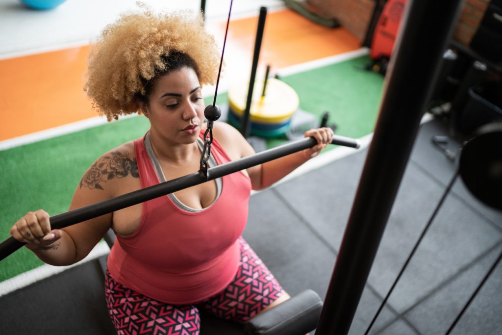 Uma mulher plus-size treinando com dedicação e disposição em uma máquina de puxada na academia, exemplificando como a perda de peso melhora a vitalidade e a disposição para atividades físicas.