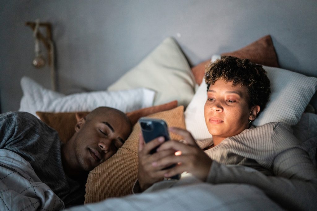 Mulher deitada na cama em um quarto escuro sofrendo de insônia, com o rosto iluminado pela tela do celular que está usando. Ao lado dela, um homem dorme tranquilamente.