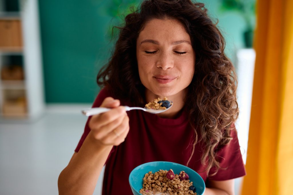 Mulher de cabelos cacheados sorrindo de olhos fechados enquanto come uma colherada de uma tigela saudável com cereais, aveia e frutas vermelhas, ilustrando a dieta para o controle da dislipidemia.