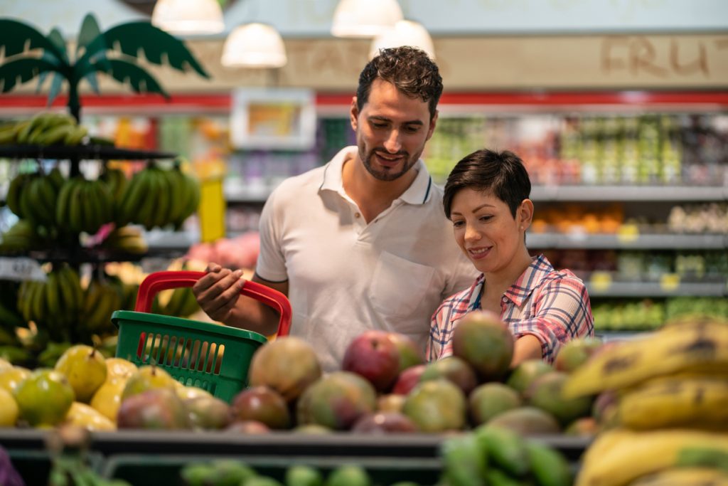 Casal sorridente na seção de hortifrúti do supermercado, segurando uma cesta de compras enquanto escolhe frutas frescas, ilustrando a prática de descascar mais e desembalar menos na reeducação alimentar.