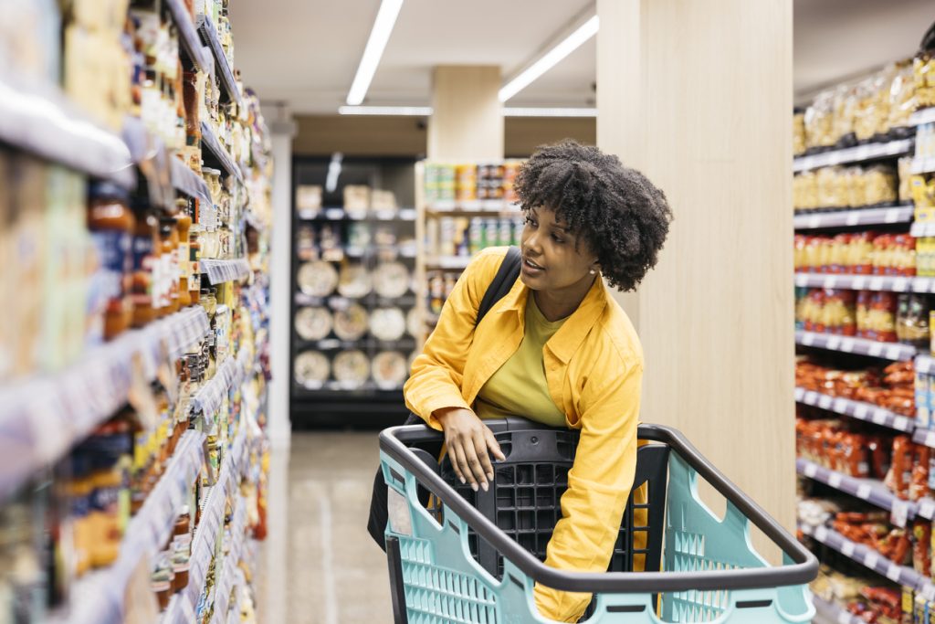 Mulher no corredor de um supermercado, apoiada em um carrinho de compras, olhando atentamente para os produtos na prateleira para entender seus gatilhos e fazer melhores escolhas.