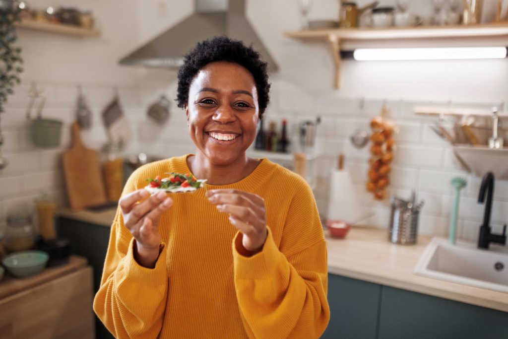 Mulher sorridente na cozinha segurando uma torrada com vegetais frescos, ilustrando ideias de lanches saudáveis para o dia a dia