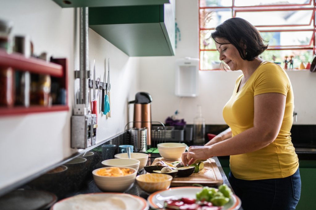 Mulher preparando alimentos na cozinha com tigelas e ingredientes sobre a bancada, ilustrando o lanche para levar ao trabalho