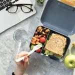 Marmita com sanduíche integral, castanhas, tomate e frutas ao lado de laptop, ilustrando lanches saudáveis que auxiliam no emagrecimento