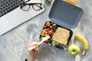 Marmita com sanduíche integral, castanhas, tomate e frutas ao lado de laptop, ilustrando lanches saudáveis que auxiliam no emagrecimento