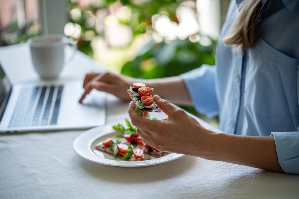 Mulher trabalhando no laptop enquanto come uma torrada integral com tomate e vegetais, mostrando opções de lanches saudáveis no emagrecimento