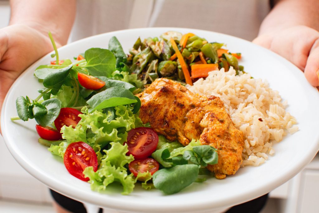 Pessoa segurando um prato branco com uma refeição saudável e colorida, contendo frango grelhado, arroz integral, salada verde com tomate cereja e legumes, representando o início de uma reeducação alimentar.