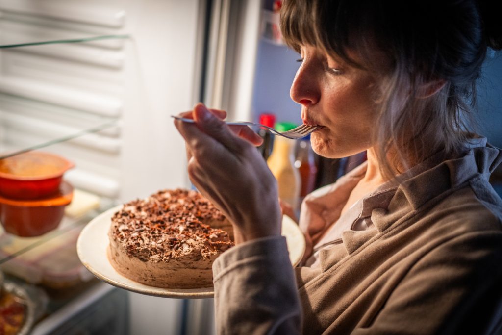 Mulher comendo bolo de chocolate em frente à geladeira aberta, representando como os hábitos alimentares afetam os resultados da caneta para emagrecer.